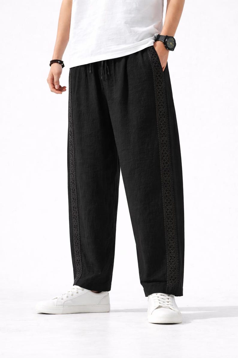 May include: Black linen pants with a drawstring waist and decorative trim along the sides. The pants are loose-fitting and paired with white sneakers. The overall style is casual and comfortable.