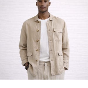 May include: A beige linen jacket with four front pockets and button fastenings. The jacket is worn over a white t-shirt and paired with matching beige trousers. The outfit is a neutral colour and appears to be made of a lightweight fabric.