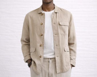 Op de afbeelding: Een beige linnen jasje met vier voorzakken en knoopsluitingen. De jas wordt gedragen over een wit t-shirt en gecombineerd met een bijpassende beige broek. De outfit is neutraal van kleur en lijkt gemaakt van een lichte stof.