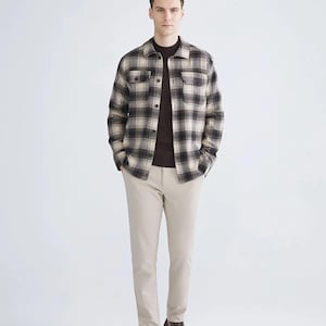 May include: A man in a brown sweater, a black, grey, and beige plaid shirt, beige trousers, and brown leather boots. The outfit is a casual, layered ensemble, suitable for cooler weather.