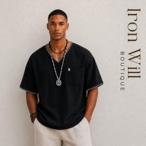 May include: Black V-neck shirt with white stitching on the collar and sleeves. The shirt has a pocket on the chest and a small logo. The model is wearing multiple necklaces and bracelets. The text "Iron Will Boutique" is on the right side.