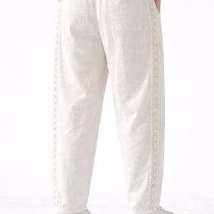 May include: Off-white linen pants with a relaxed fit. The pants feature a decorative embroidered trim along the outer seams and a single back pocket. The waistband is elasticized. The pants are paired with white sneakers.