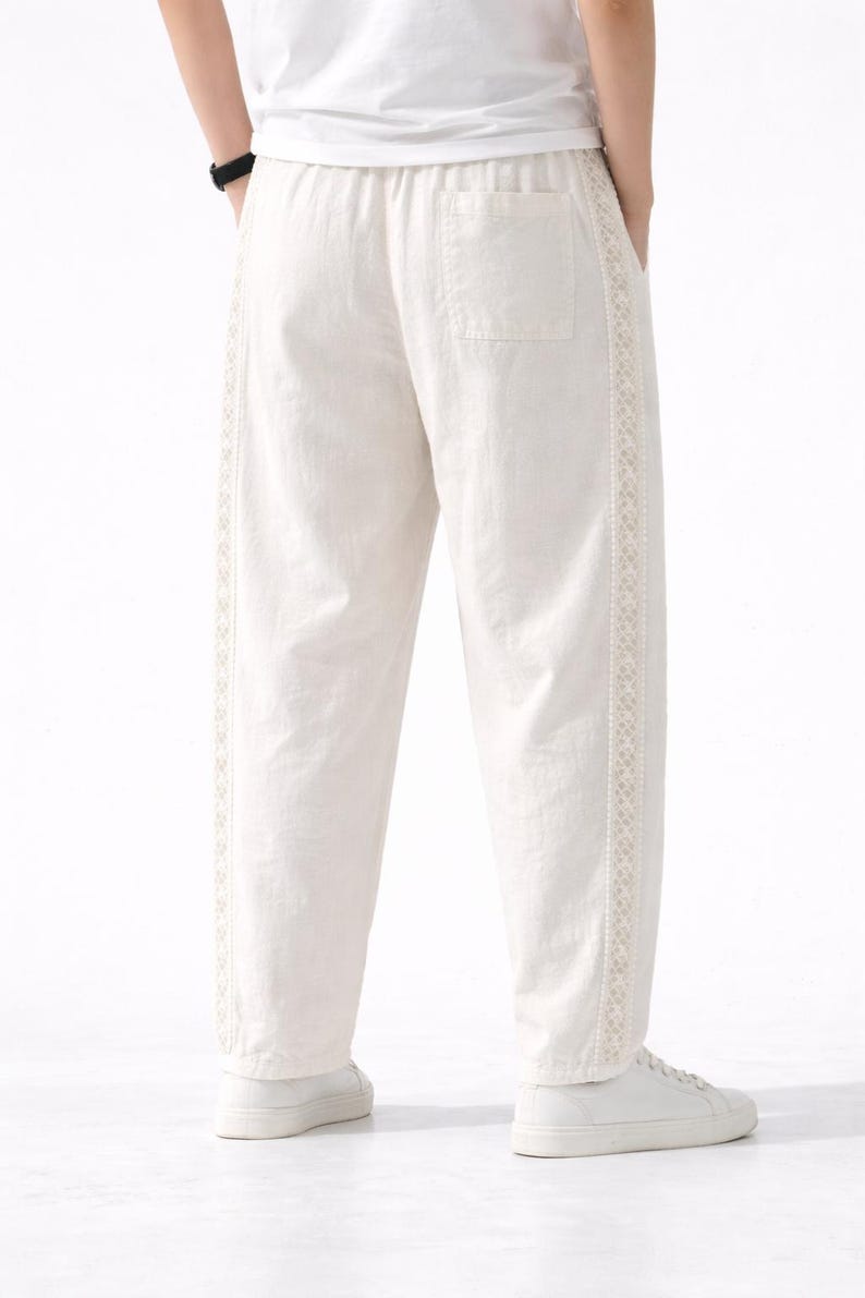 May include: Off-white linen pants with a relaxed fit. The pants feature a decorative embroidered trim along the outer seams and a single back pocket. The waistband is elasticized. The pants are paired with white sneakers.