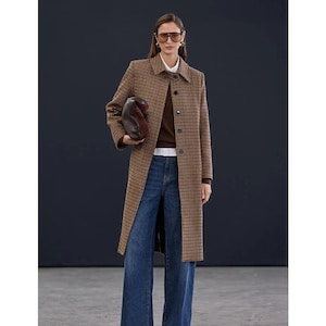 Puede incluir: Una mujer con un abrigo a cuadros marrón y negro, un suéter marrón y una camisa blanca, con vaqueros azules. Sostiene un bolso marrón y lleva gafas de sol. El abrigo es largo hasta la rodilla.