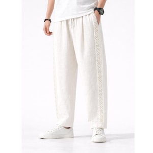 May include: Off-white linen pants with a drawstring waist and decorative trim along the side seams. The pants are paired with white sneakers. The style is casual and comfortable.