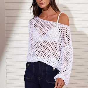 Op de afbeelding: Een witte, open geweven gehaakte top met lange mouwen en een off-the-shoulder ontwerp. De top wordt gecombineerd met een donkerblauwe jeans. Het haakpatroon cre&euml;ert een doorschijnend effect. De jeans heeft grote zakken en gouden knopen.