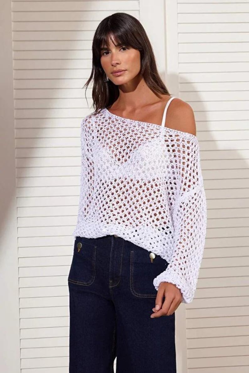 Op de afbeelding: Een witte, open geweven gehaakte top met lange mouwen en een off-the-shoulder ontwerp. De top wordt gecombineerd met een donkerblauwe jeans. Het haakpatroon cre&euml;ert een doorschijnend effect. De jeans heeft grote zakken en gouden knopen.