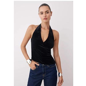 Op de afbeelding: Een zwarte haltertop met een diepe V-hals. De top is gemaakt van een zacht, fluweelachtig materiaal. De persoon draagt een donkerblauwe jeans en zilveren armbanden.