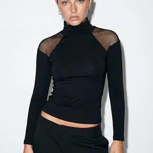 May include: A black long-sleeved turtleneck top with mesh detailing on the shoulders, worn with black trousers. The mesh adds a textural contrast to the smooth fabric. A simple, modern outfit in black.