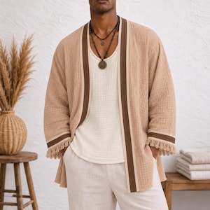 May include: A beige textured open-front kimono with brown and cream striped trim and fringe at the sleeves. The garment is worn over a cream-colored tank top and light-colored trousers. The text Iron Will BOUTIQUE appears in the upper right corner.