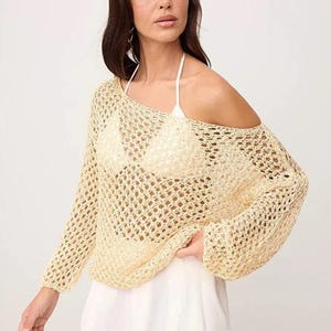 Op de afbeelding: Een lichtgele, open gebreide trui met lange mouwen en een off-the-shoulder ontwerp. De trui wordt gecombineerd met een witte broek en een witte haltertop. Het breiwerk heeft een losse, luchtige textuur, perfect voor warm weer.