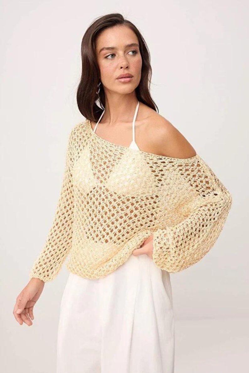 Op de afbeelding: Een lichtgele, open gebreide trui met lange mouwen en een off-the-shoulder ontwerp. De trui wordt gecombineerd met een witte broek en een witte haltertop. Het breiwerk heeft een losse, luchtige textuur, perfect voor warm weer.