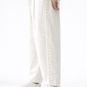 May include: White linen pants with a decorative embroidered stripe down the side. The pants are paired with white sneakers. The drawstring waist pants are a casual style.
