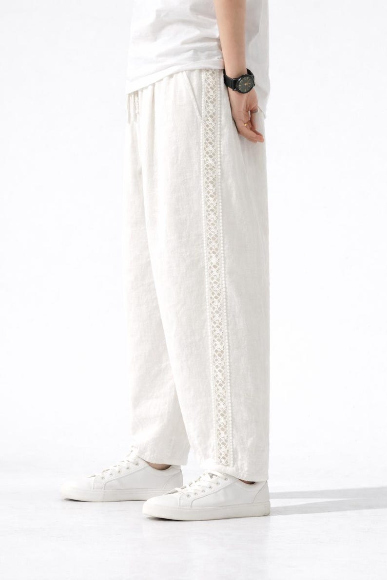May include: White linen pants with a decorative embroidered stripe down the side. The pants are paired with white sneakers. The drawstring waist pants are a casual style.