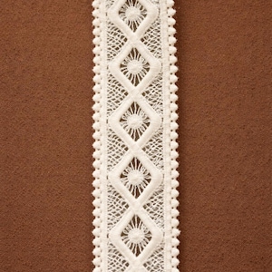May include: Close-up of a white lace trim with a repeating diamond and floral pattern. The lace has a scalloped edge and is set against a brown background. Ideal for sewing and craft projects.