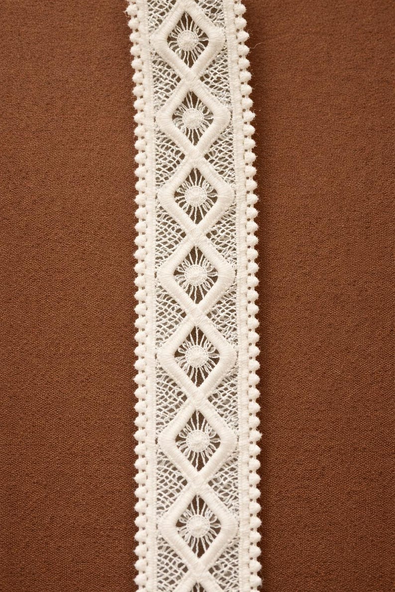 May include: Close-up of a white lace trim with a repeating diamond and floral pattern. The lace has a scalloped edge and is set against a brown background. Ideal for sewing and craft projects.