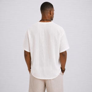 May include: A white short-sleeved shirt with a textured appearance and a rounded hemline. The shirt has a V-neck collar and is paired with light beige pants. The model is wearing a beaded necklace and bracelet.