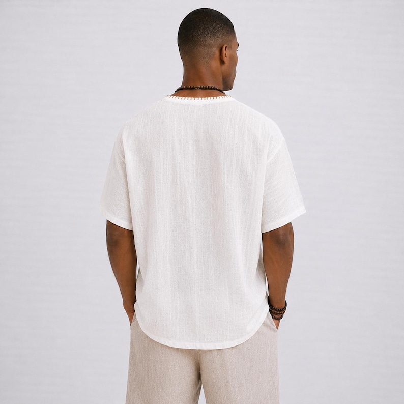 May include: A white short-sleeved shirt with a textured appearance and a rounded hemline. The shirt has a V-neck collar and is paired with light beige pants. The model is wearing a beaded necklace and bracelet.