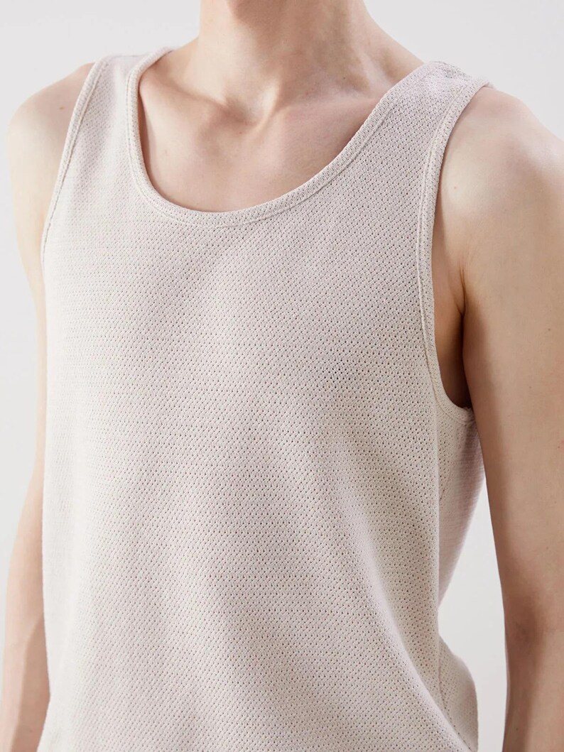 May include: A light beige tank top with a scoop neckline. The sleeveless shirt has a textured, perforated knit pattern. The image shows the top from the chest up, with the shoulder and arm visible.