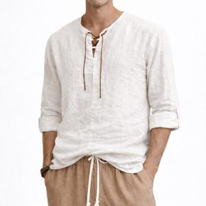 May include: A white linen shirt with a lace-up neckline and long sleeves, worn with brown drawstring trousers. The shirt has rolled-up sleeves and the trousers have a white drawstring. The outfit is casual and comfortable.