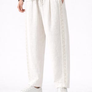 May include: Off-white linen pants with a drawstring waist and decorative lace trim down the sides. The pants are paired with white sneakers and a white t-shirt. The overall style is casual and comfortable.