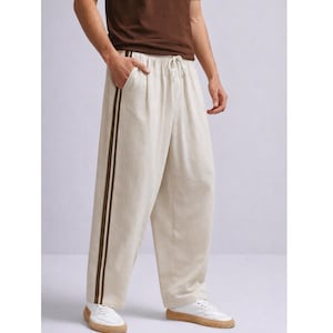 Puede incluir: Pantalones de lino color crema con cintura con cord&oacute;n y dos rayas marr&oacute;n oscuro en el lateral. Los pantalones de pernera ancha se llevan con una camiseta marr&oacute;n y zapatillas blancas.