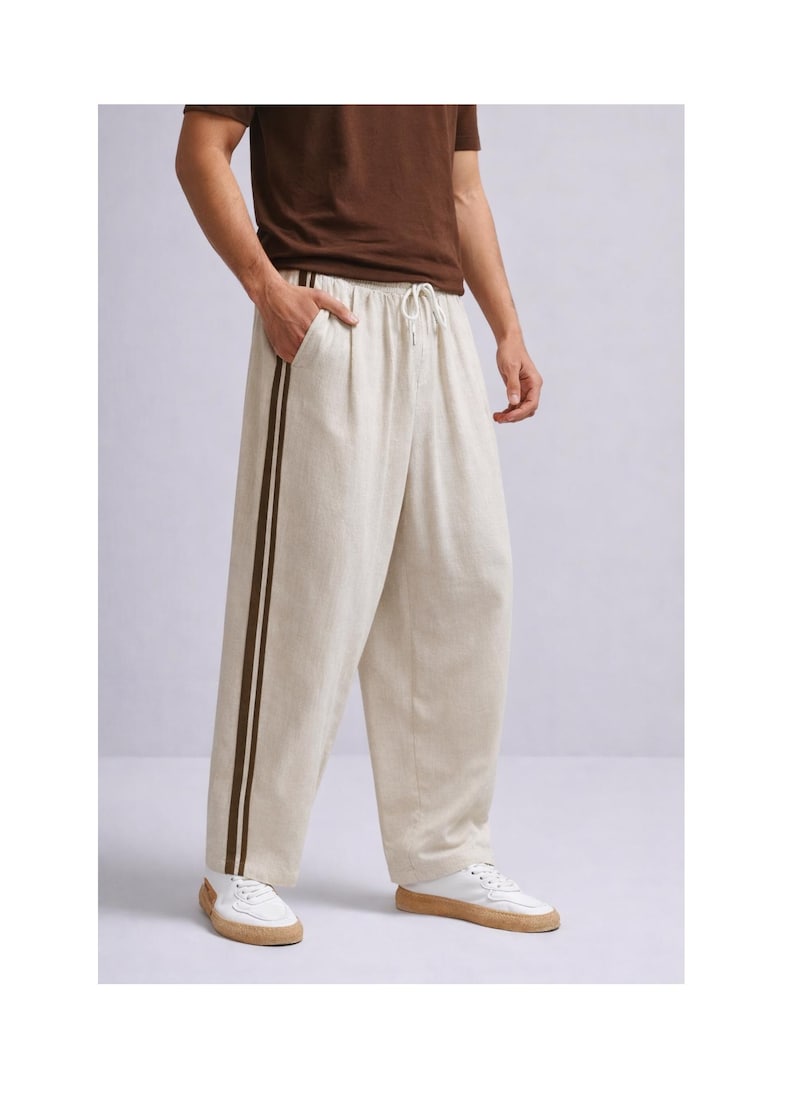 Puede incluir: Pantalones de lino color crema con cintura con cord&oacute;n y dos rayas marr&oacute;n oscuro en el lateral. Los pantalones de pernera ancha se llevan con una camiseta marr&oacute;n y zapatillas blancas.