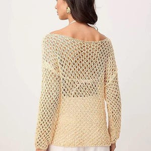 Op de afbeelding: Een lichtbeige, opengebreide gehaakte trui met lange mouwen. De trui heeft een boothals en een losse, relaxte pasvorm. Het model draagt een witte broek. De trui is een stijlvol, lichtgewicht kledingstuk, perfect om te layeren.