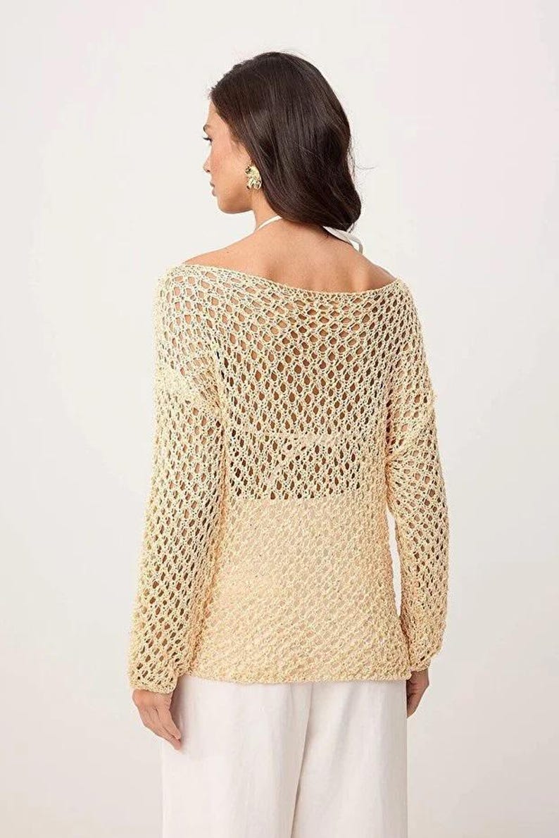 Op de afbeelding: Een lichtbeige, opengebreide gehaakte trui met lange mouwen. De trui heeft een boothals en een losse, relaxte pasvorm. Het model draagt een witte broek. De trui is een stijlvol, lichtgewicht kledingstuk, perfect om te layeren.