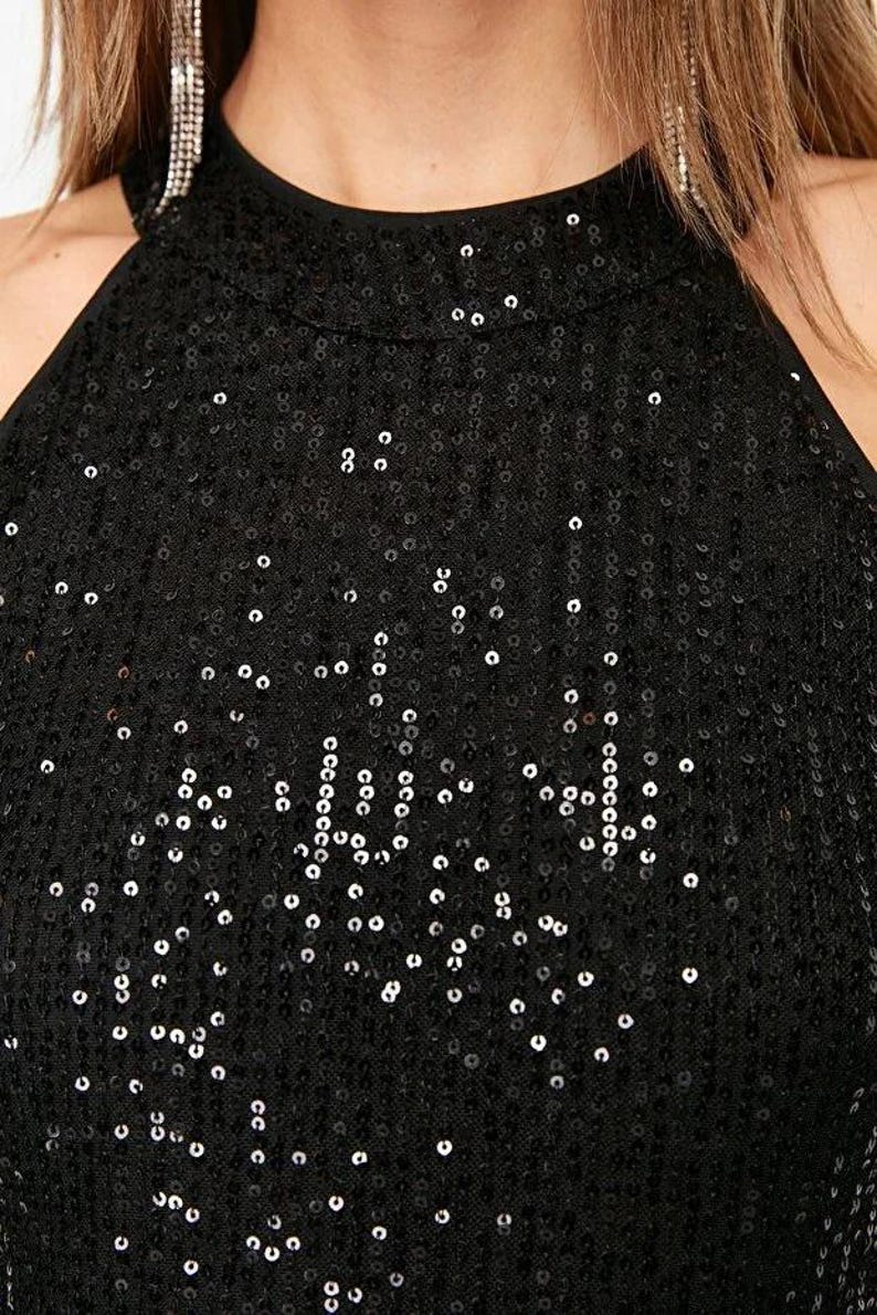 May include: Close-up of a black sequin halter top. The top features a high neckline and is covered in small, reflective sequins. The garment is likely part of a formal wear collection, suitable for evening events or parties.
