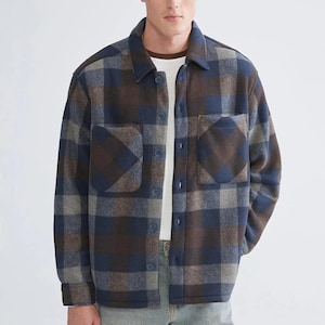 Puede incluir: Chaqueta de camisa a cuadros en tonos marrón, azul y gris con cierre de botones y dos bolsillos delanteros. La chaqueta tiene cuello camisero. El modelo lleva una camiseta blanca debajo. La chaqueta está hecha de un material grueso y cálido.