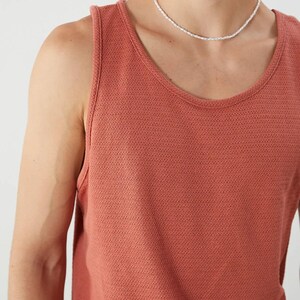 May include: A coral-colored tank top with a scoop neck. The tank top is made of a textured knit fabric. A white beaded necklace is worn around the neck.