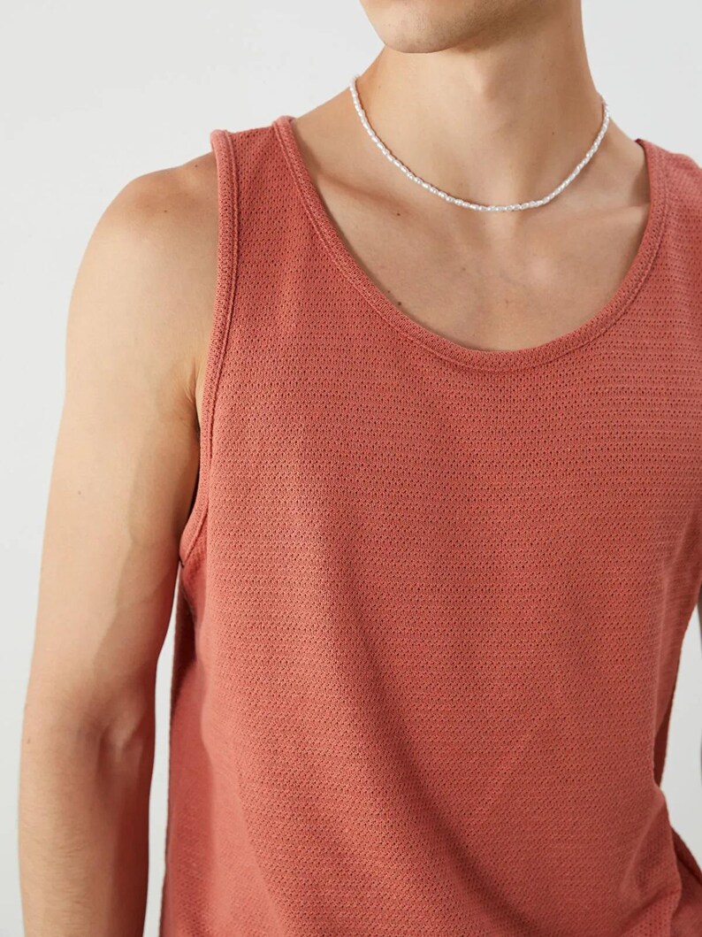 May include: A coral-colored tank top with a scoop neck. The tank top is made of a textured knit fabric. A white beaded necklace is worn around the neck.