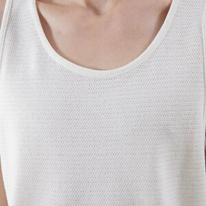May include: Close-up of a white tank top with a scoop neckline. The fabric has a textured, perforated pattern. The top is sleeveless and appears to be made of a lightweight material, suitable for warm weather.