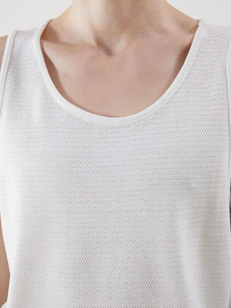 May include: Close-up of a white tank top with a scoop neckline. The fabric has a textured, perforated pattern. The top is sleeveless and appears to be made of a lightweight material, suitable for warm weather.