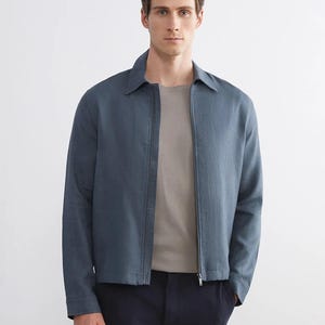 Puede incluir: Chaqueta azul con cierre de cremallera y cuello camisero. Se combina con una camiseta gris claro y pantalones azul oscuro. El conjunto es sencillo y moderno, adecuado para ropa informal.
