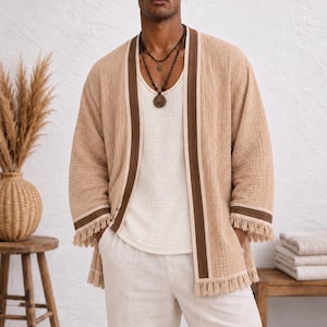 May include: A beige open-front kimono jacket featuring dark brown trim along the lapels and cuffs, finished with a fringe hem. The garment is worn over a white tank top and light-colored trousers. The text Iron Will BOUTIQUE is visible.