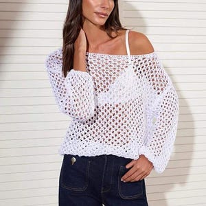 Op de afbeelding: Witte gehaakte top met lange mouwen en een off-the-shoulder ontwerp. De top wordt gecombineerd met een donkerblauwe jeans. De open structuur van de top cre&euml;ert een luchtige, zomerse look. De outfit is eenvoudig en stijlvol.