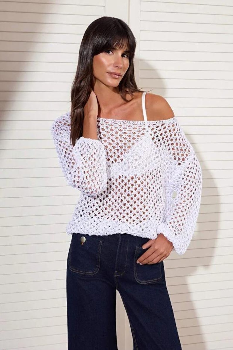 Op de afbeelding: Witte gehaakte top met lange mouwen en een off-the-shoulder ontwerp. De top wordt gecombineerd met een donkerblauwe jeans. De open structuur van de top cre&euml;ert een luchtige, zomerse look. De outfit is eenvoudig en stijlvol.