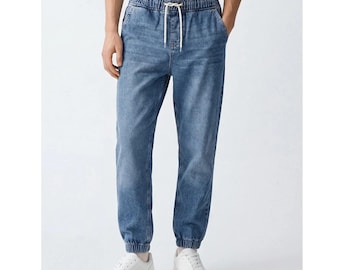 May include: Blue denim joggers featuring a light wash finish, an elasticized waistband with a white drawstring, and elastic cuffs at the ankles. The pants have a relaxed fit through the legs and are paired with plain white sneakers.