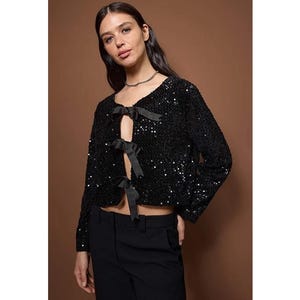 May include: A black sequined jacket with long sleeves and a V-neckline. The jacket features three black satin bows down the front. The model is wearing black pants and a silver necklace.