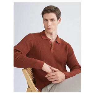 Puede incluir: Un polo de manga larga de color óxido con textura acanalada. La camisa tiene un cuello clásico y una tapeta de tres botones. El modelo está sentado en una silla de madera, vistiendo pantalones de color claro.