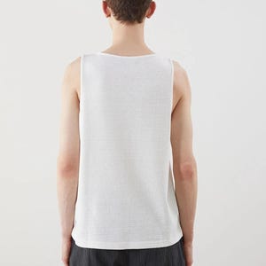 May include: A white, textured tank top. The sleeveless shirt has a square neckline and a subtle, woven pattern. The person is wearing dark gray pants. The background is plain white.
