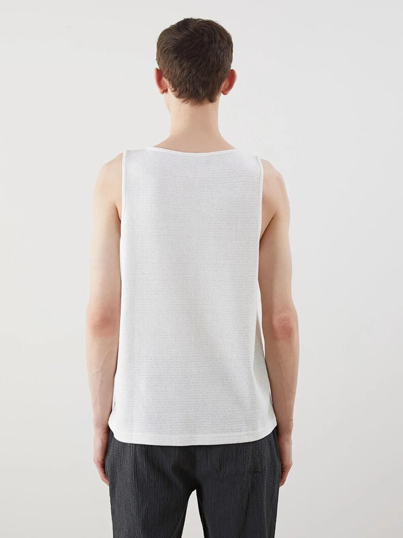 May include: A white, textured tank top. The sleeveless shirt has a square neckline and a subtle, woven pattern. The person is wearing dark gray pants. The background is plain white.