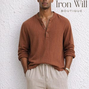 Puede incluir: Un hombre con una camisa de manga larga color óxido con cuello Henley y pantalones beige con cordón. El texto "Iron Will Boutique" es visible. El modelo lleva un reloj y un collar dorados. Ropa informal.