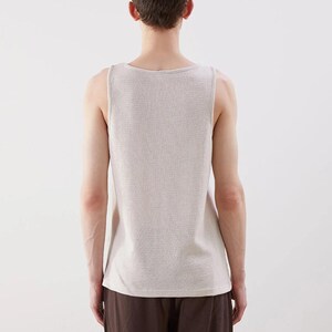 May include: A light beige tank top with a textured knit pattern. The sleeveless top has a scoop neckline and is paired with brown pants. The image is taken from the back, showcasing the clothing's design and fit.