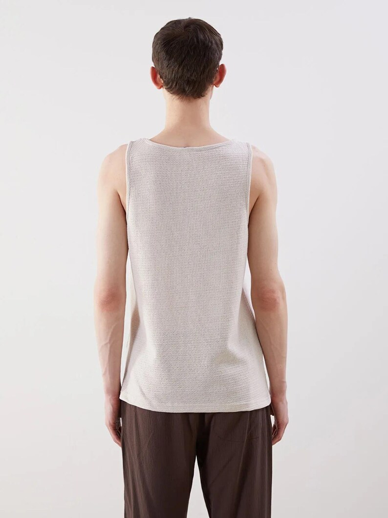 May include: A light beige tank top with a textured knit pattern. The sleeveless top has a scoop neckline and is paired with brown pants. The image is taken from the back, showcasing the clothing's design and fit.