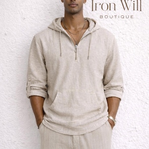 May include: A beige linen hoodie with a front pocket and a quarter-zip closure, paired with matching linen pants. The outfit is from Iron Will Boutique. The model is wearing a watch and a necklace.