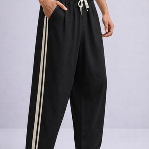 Puede incluir: Pantalones negros de pernera ancha con dos rayas verticales de color crema en el lateral. Los pantalones tienen cintura con cord&oacute;n y se combinan con zapatillas blancas. El modelo lleva una camiseta blanca.