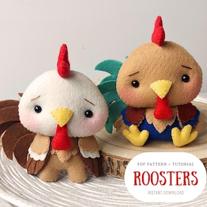 Pode incluir: Padrão PDF e tutorial para fazer dois brinquedos de galo de feltro. Um galo é marrom com detalhes azuis e o outro é branco com detalhes marrons. O texto "ROOSTERS" está impresso em vermelho em uma superfície de madeira.